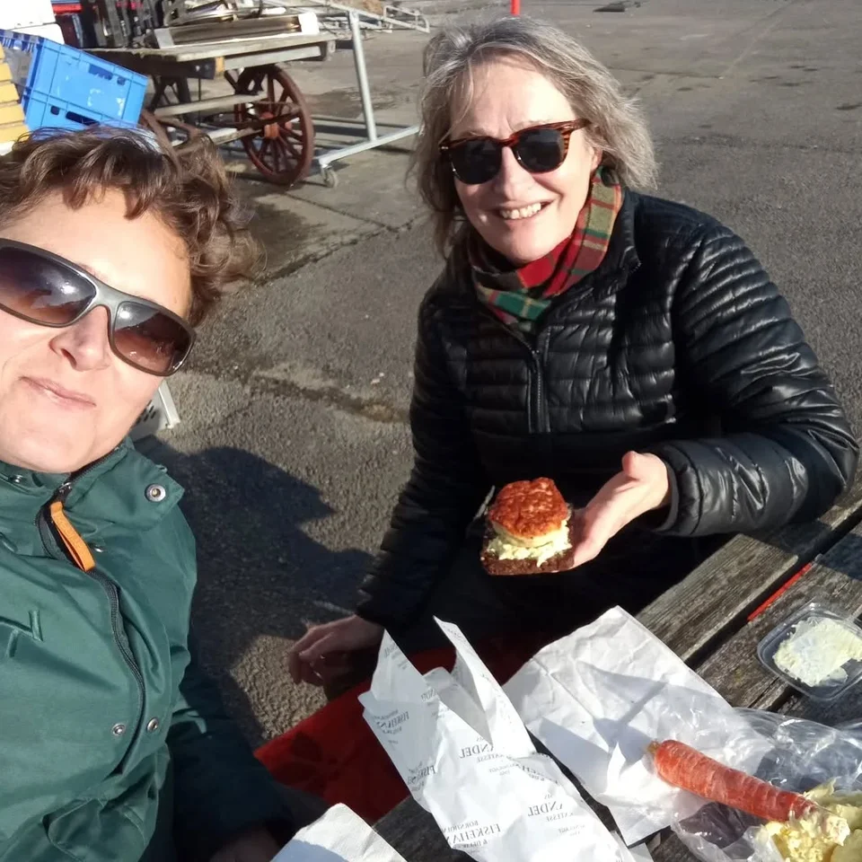 Nina Trige Andersen og Marianne Bækbøl spiser madpakke ved et bord på en havn